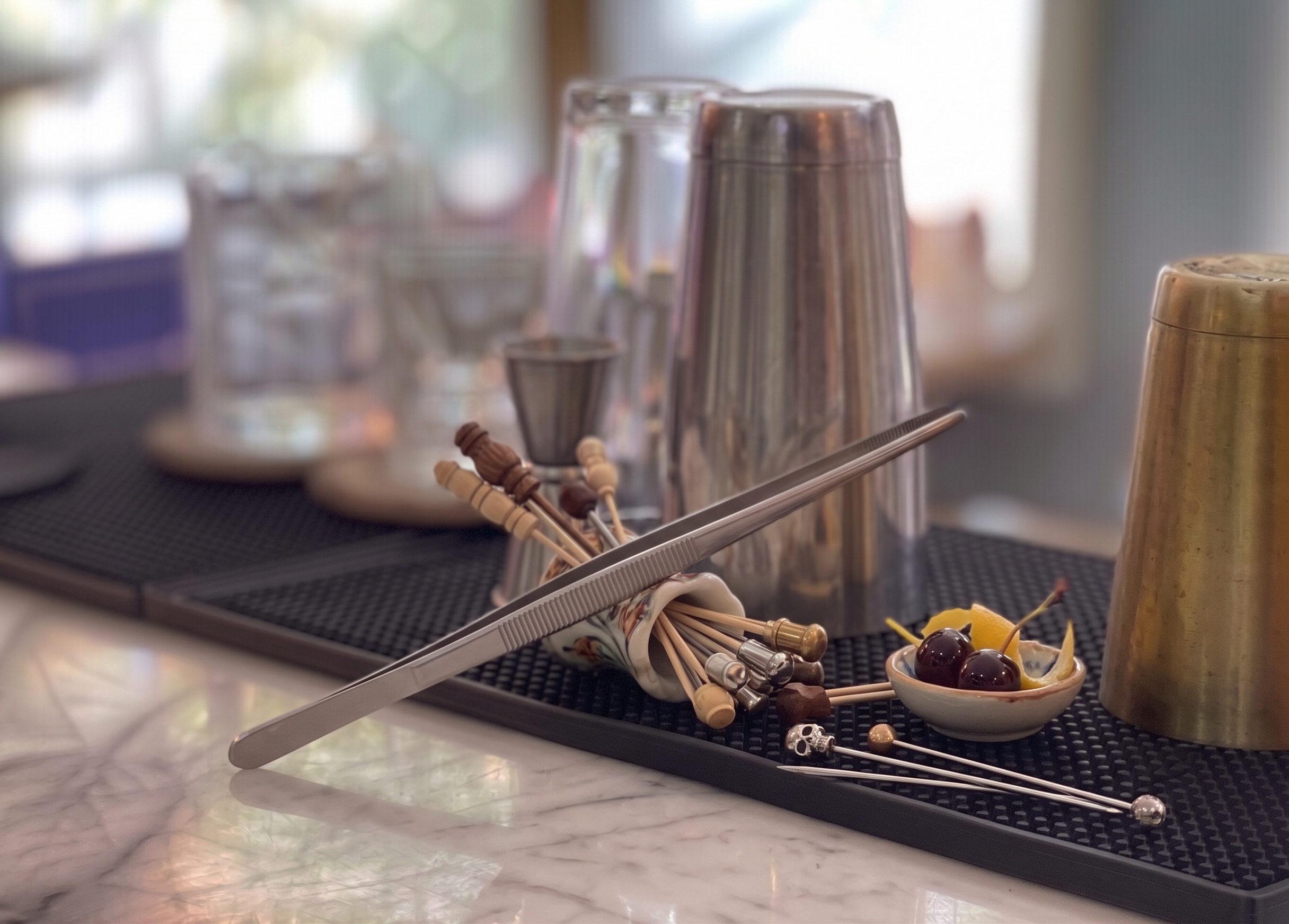 Pinzas de bar y palillos de decoración con guindas y cáscara de naranja, sobre una barra.