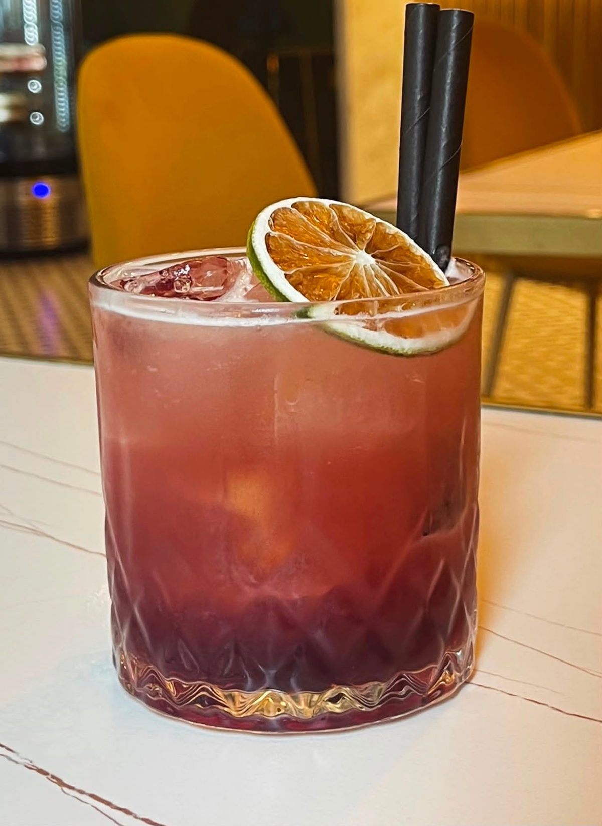Vaso Old Fashioned con Bramble, degradado púrpura, hielo y una rodaja de cítrico deshidratado.